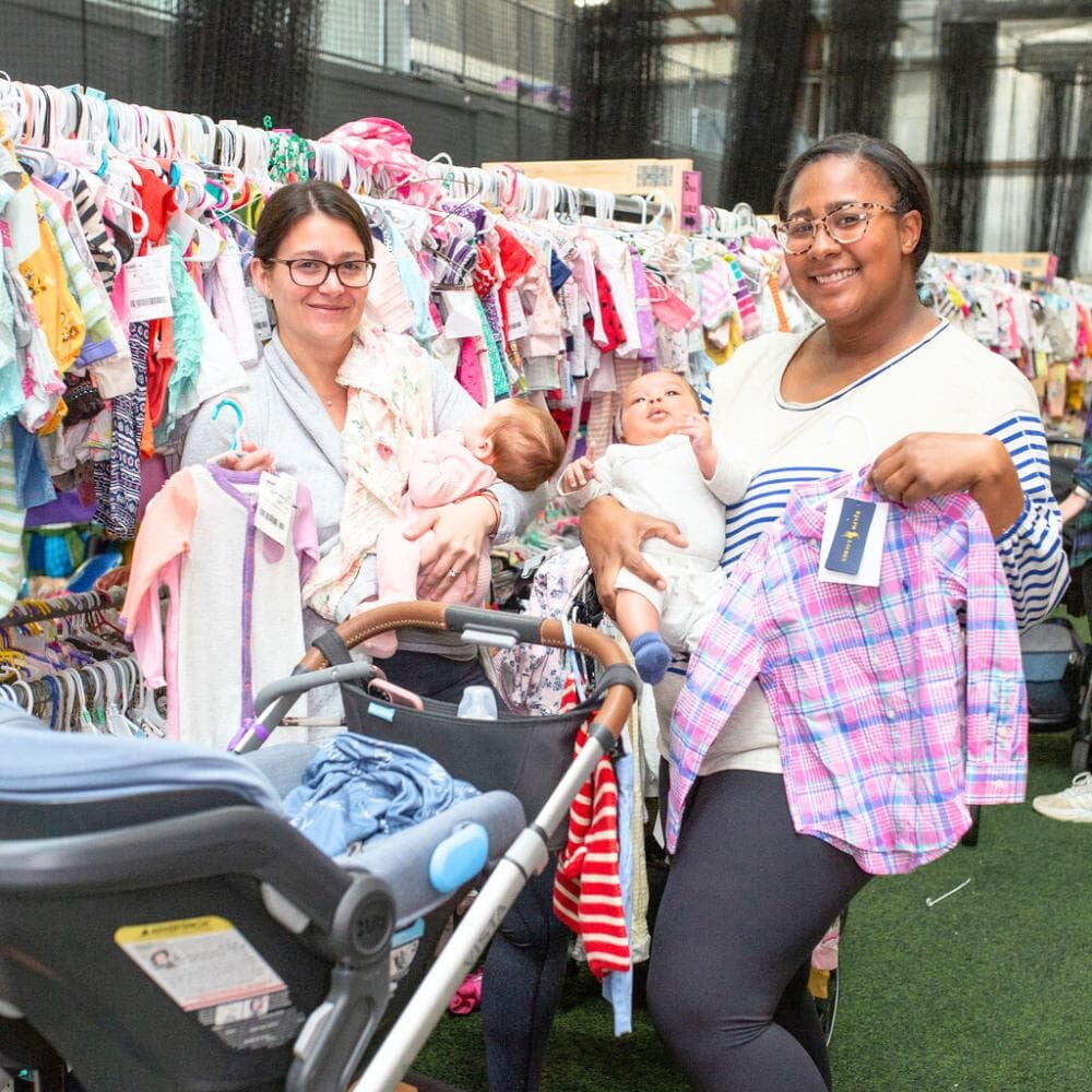 Two friends shop together at their local JBF sale. Both moms carry their baby while holding clothes.