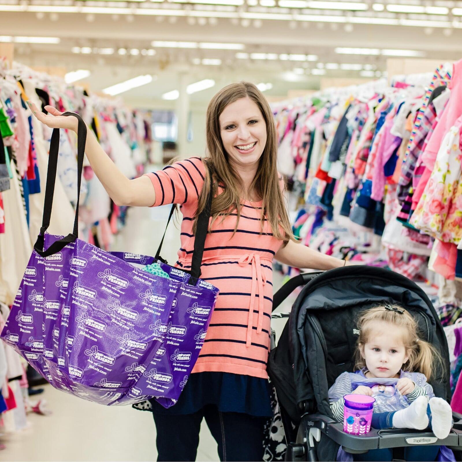 A Mom holds up her JBF shopping bag with her right hand while resting her left hand on her stroller with toddler daughter inside.